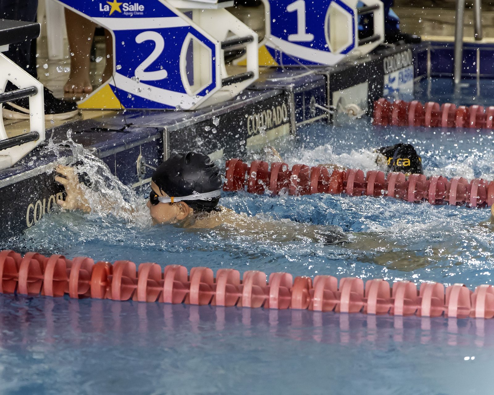 Atleta de touca preta toca a borda da piscina logo abaixo do bloco de partida da raia 2, no 1º FEN, em março de 2026, no La Salle Águas Claras