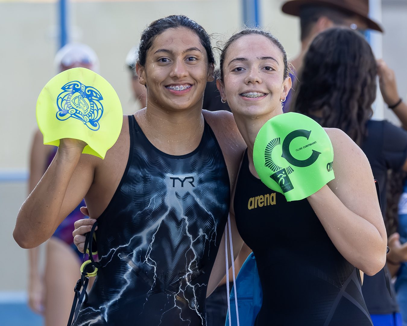 Duas atletas sorriem à beira da piscina segurando as toucas dos seus clubes