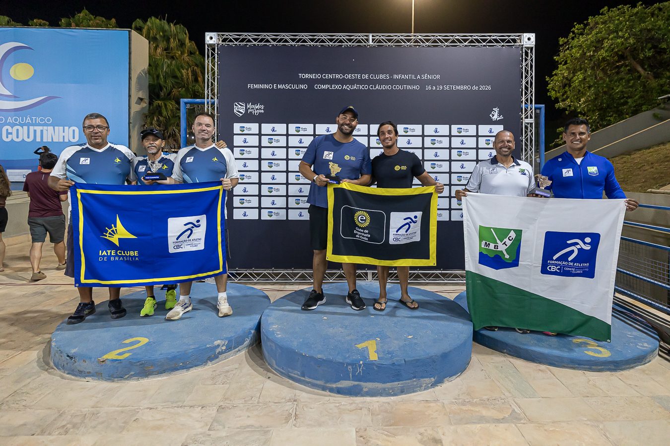 Pódio da premiação geral à noite, com três duplas de dirigentes segurando as bandeiras dos clubes: Iate Clube de Brasília no segundo lugar, Clube de Engenharia de Goiás no primeiro e MBTC no terceiro, diante do painel do torneio