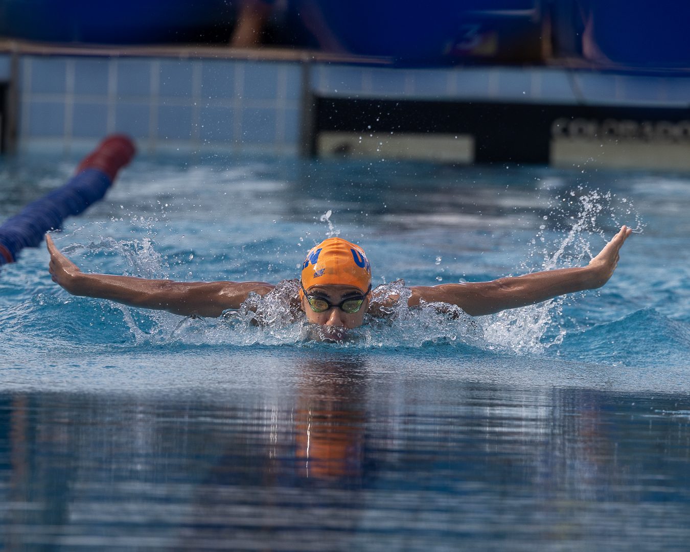 Atleta de touca laranja avança em nado borboleta com os dois braços abertos sobre a água, em piscina de competição