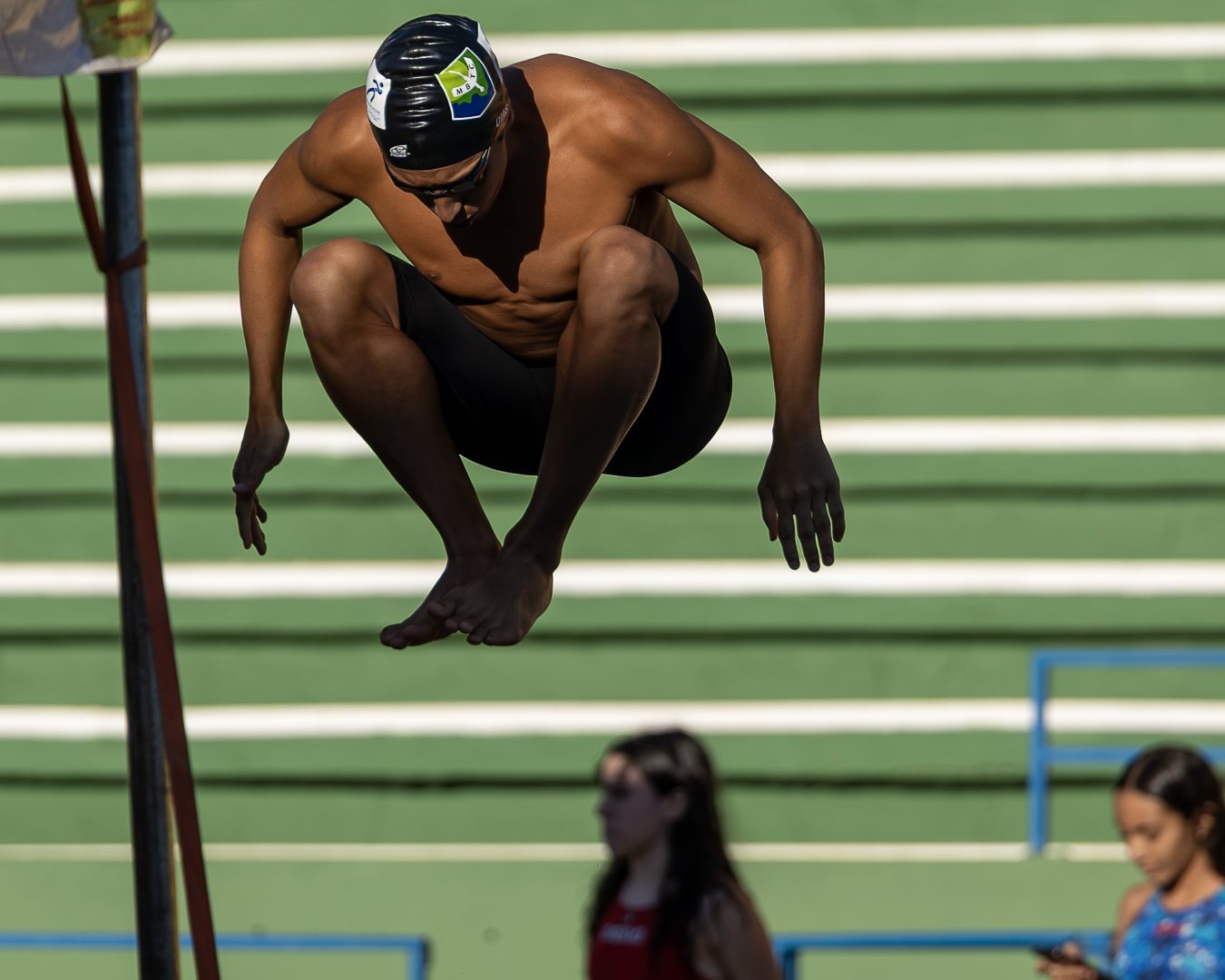 Atleta de touca preta salta no lugar, com o corpo encolhido no ar, na área de espera, diante da arquibancada
