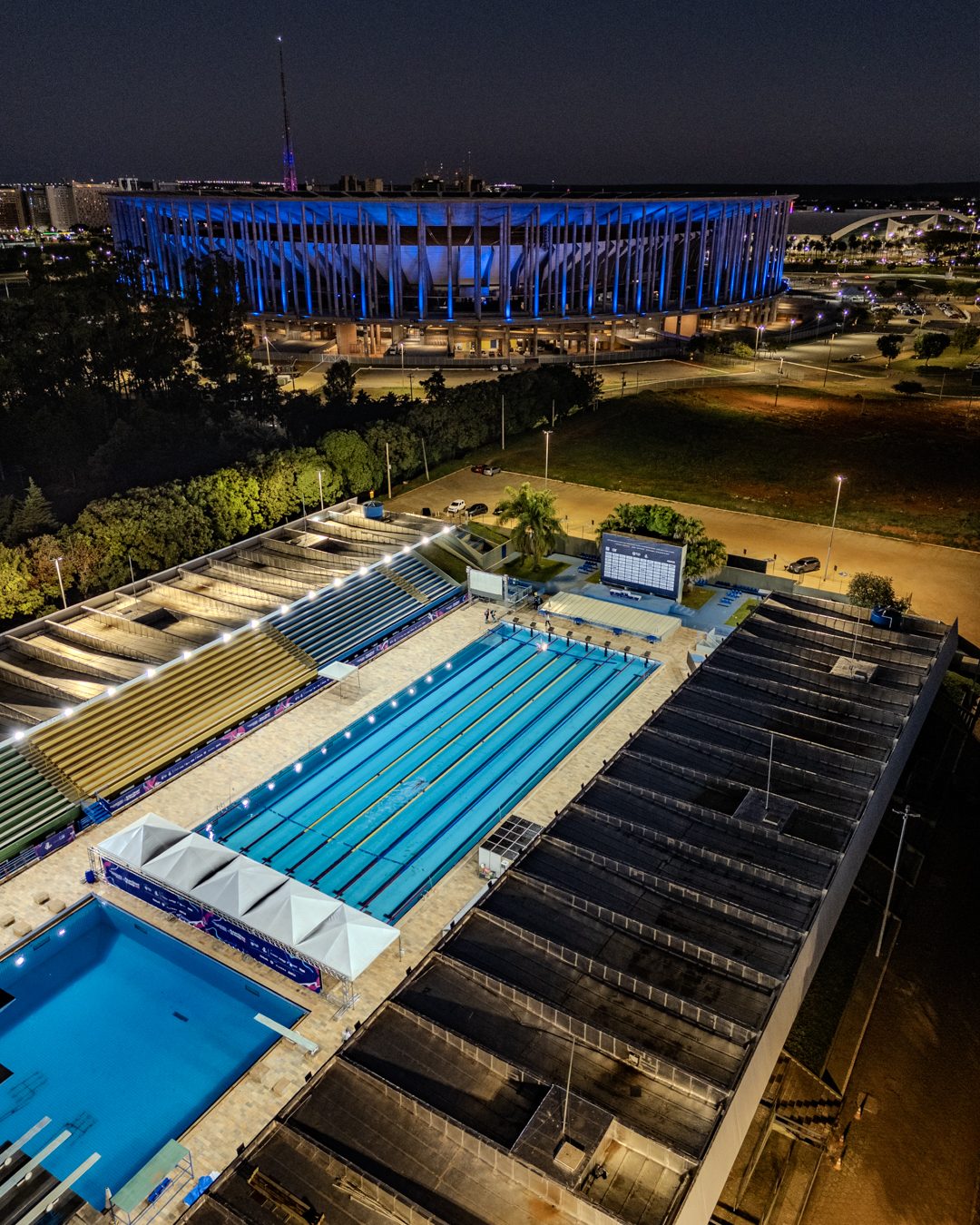 Vista aérea noturna do Complexo Aquático Cláudio Coutinho, em Brasília, com a piscina de 50 metros iluminada e o Estádio Mané Garrincha ao fundo