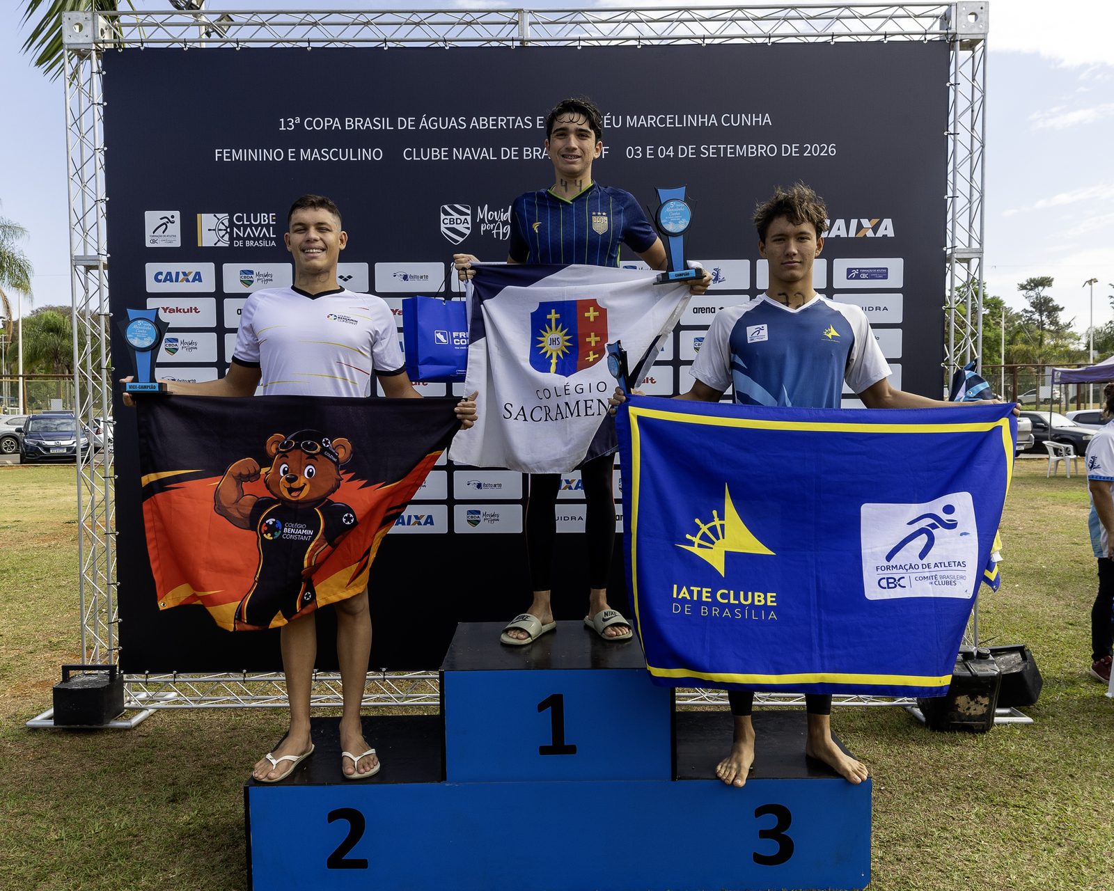 Pódio masculino dos 2,5 km, com os três medalhistas segurando troféus e bandeiras dos clubes
