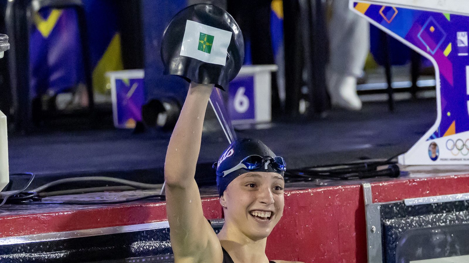 Atleta da Seleção do DF sorri dentro da piscina e ergue a touca com a bandeira do Distrito Federal depois da final