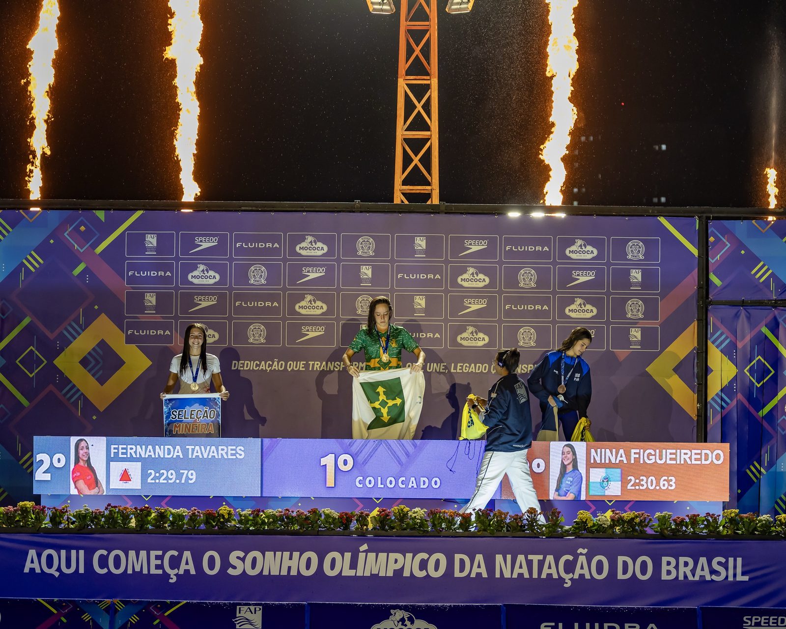 Cerimônia de premiação dos 200 metros medley infantil, com labaredas no alto do palco; a campeã segura a bandeira do Distrito Federal