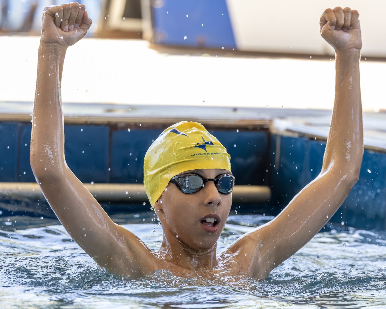 Atleta de touca amarela ergue os dois braços na piscina depois da prova