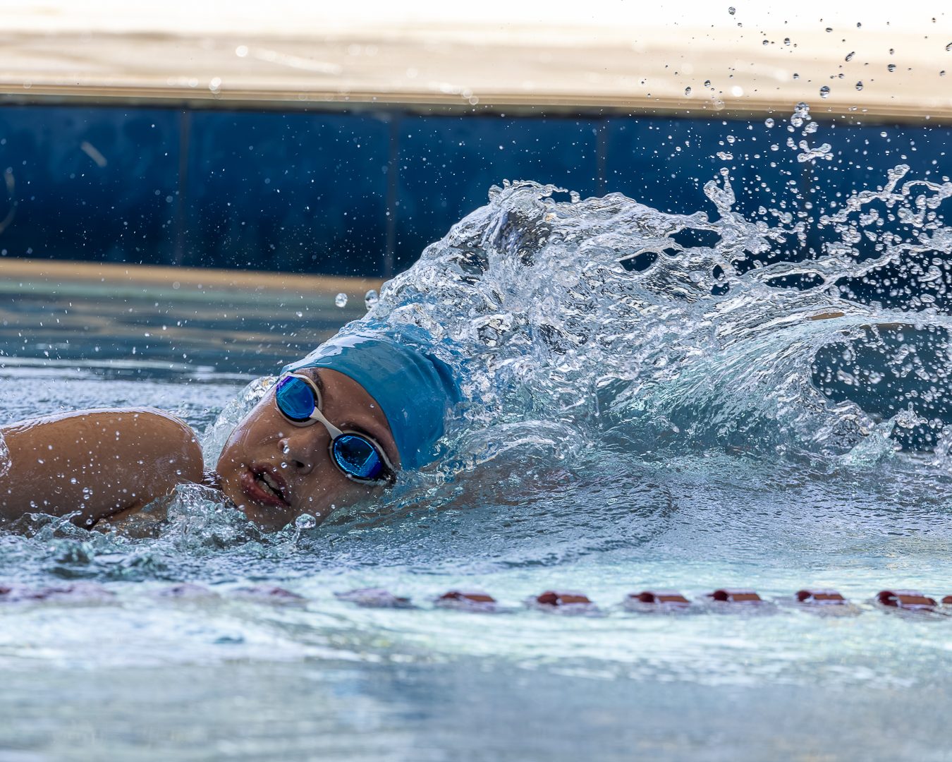 Atleta de touca azul-clara respira de lado no nado livre, entre respingos, com a raia vermelha e branca à frente