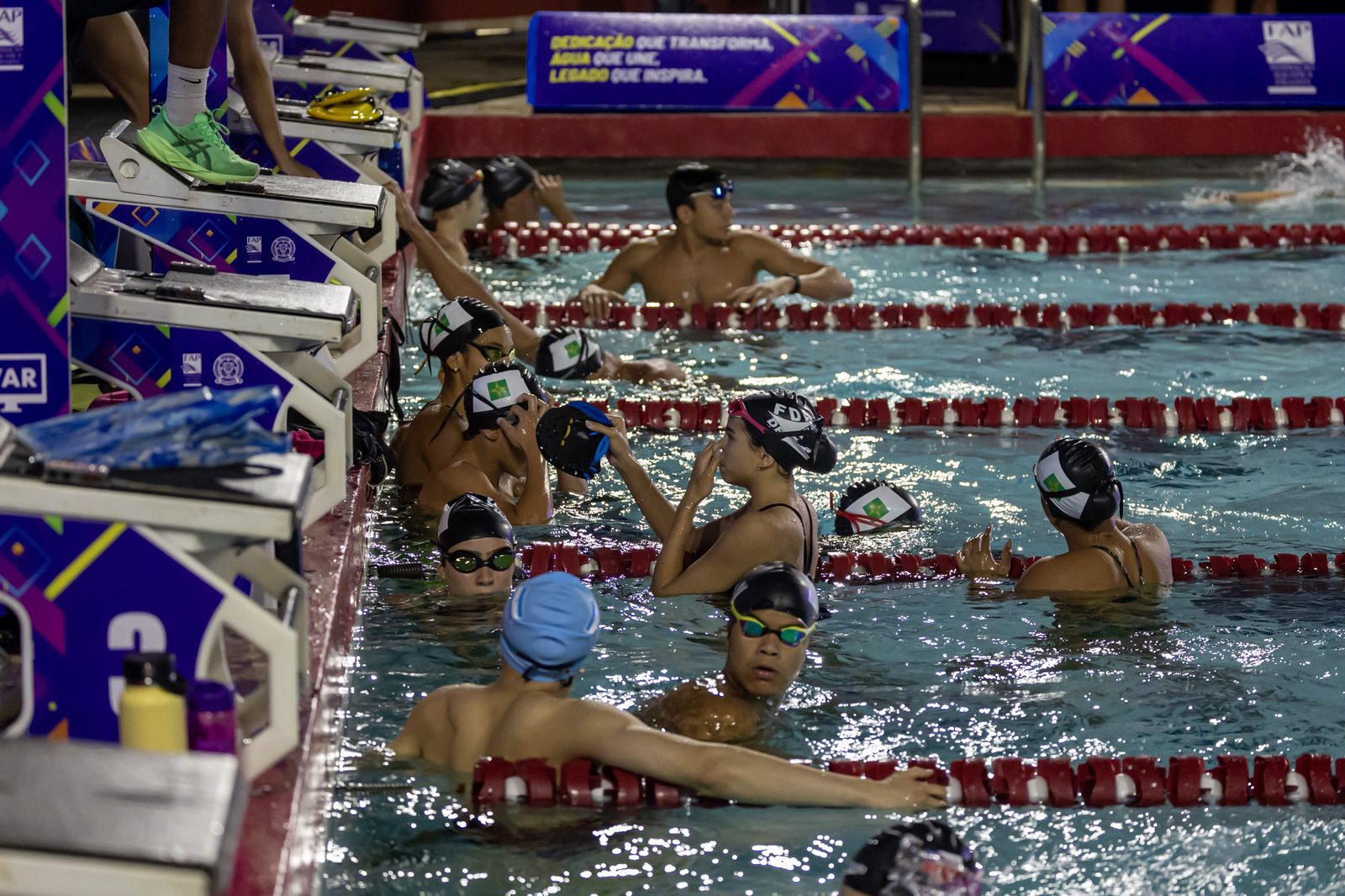 Atletas da Seleção do DF, de touca com a bandeira do Distrito Federal, reunidos na borda da piscina durante o reconhecimento