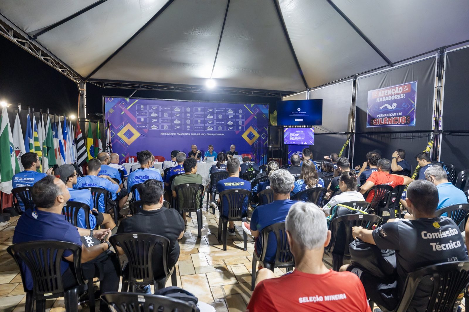 Técnicos das delegações sentados sob a tenda durante o congresso técnico, com a mesa da organização ao fundo