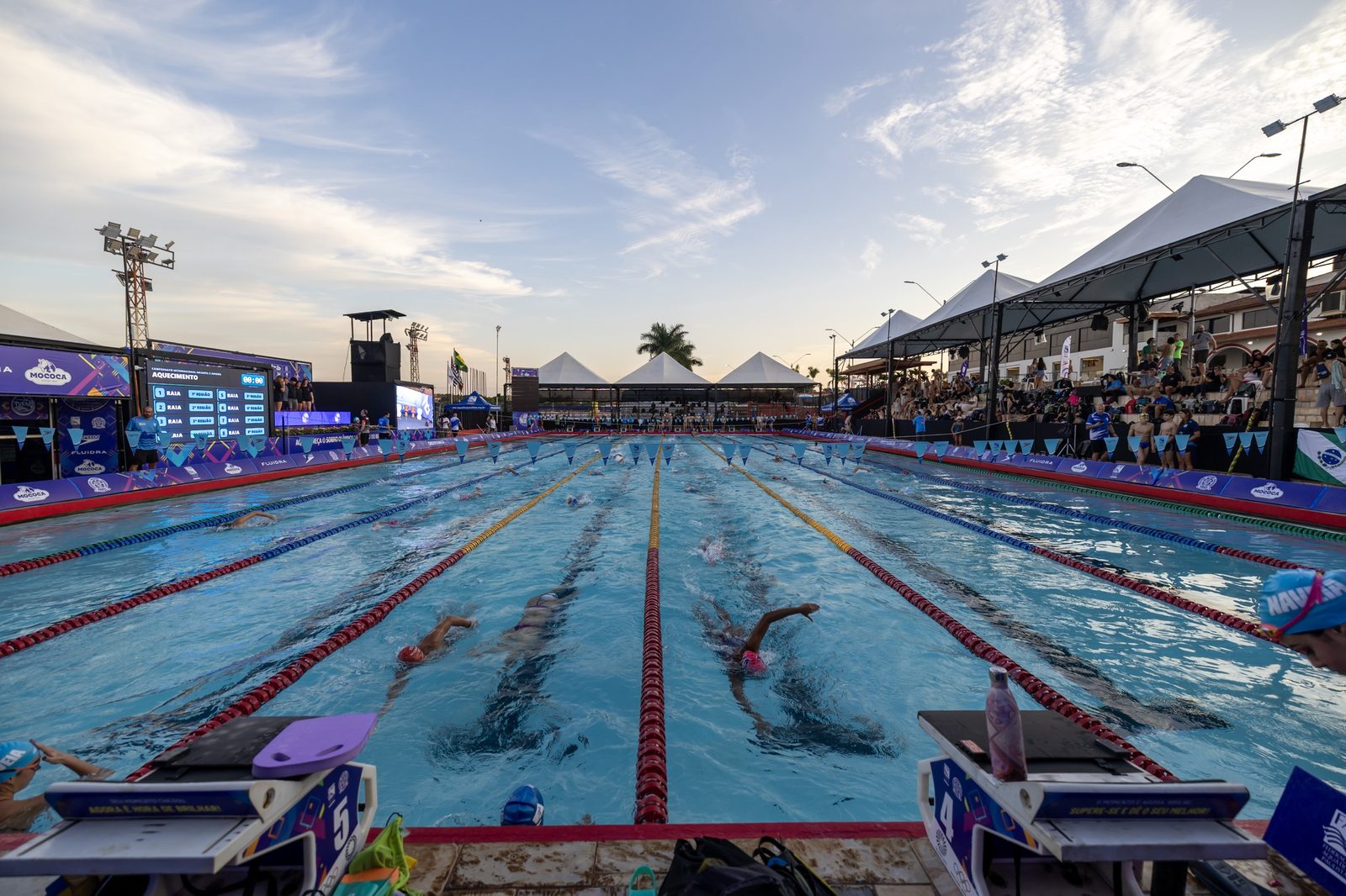Piscina de 50 metros da Associação Esportiva Mocoquense ao entardecer, com nadadores aquecendo nas raias e a arquibancada coberta à direita