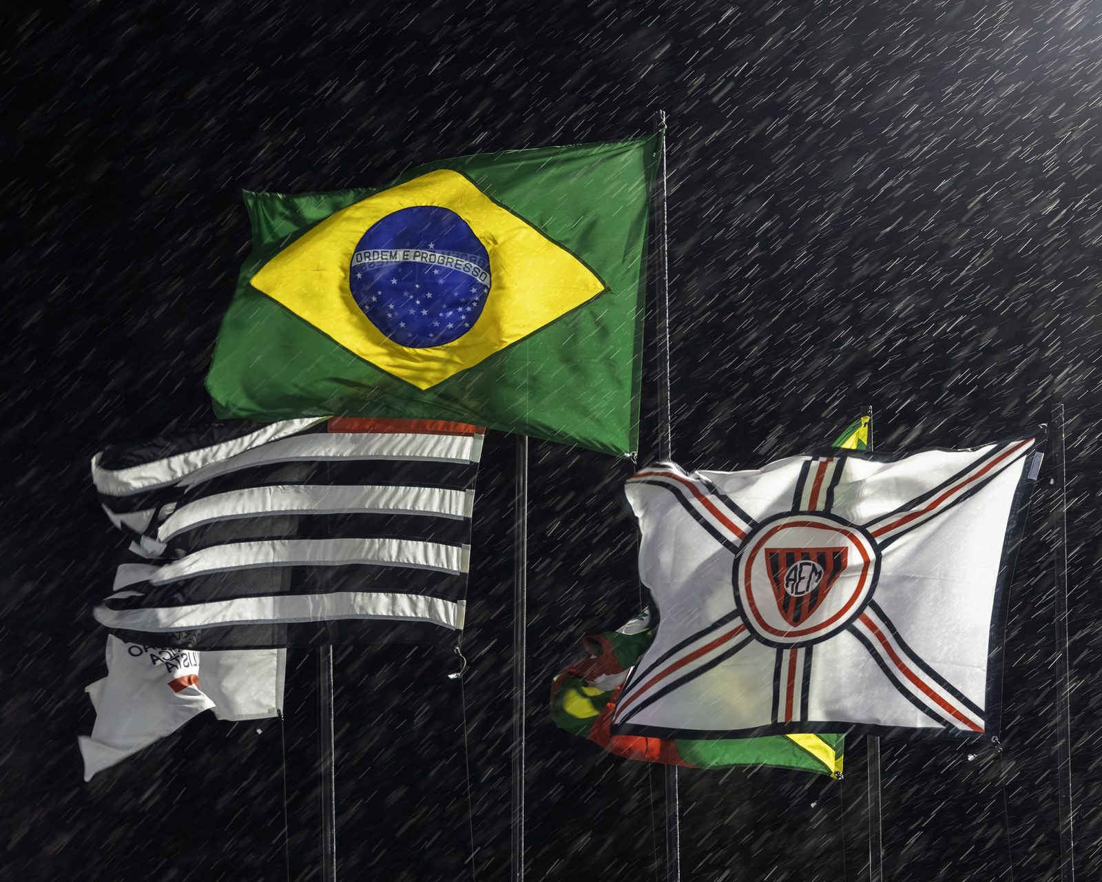 Bandeiras do Brasil, do estado de São Paulo e da Associação Esportiva Mocoquense sob a chuva, à noite