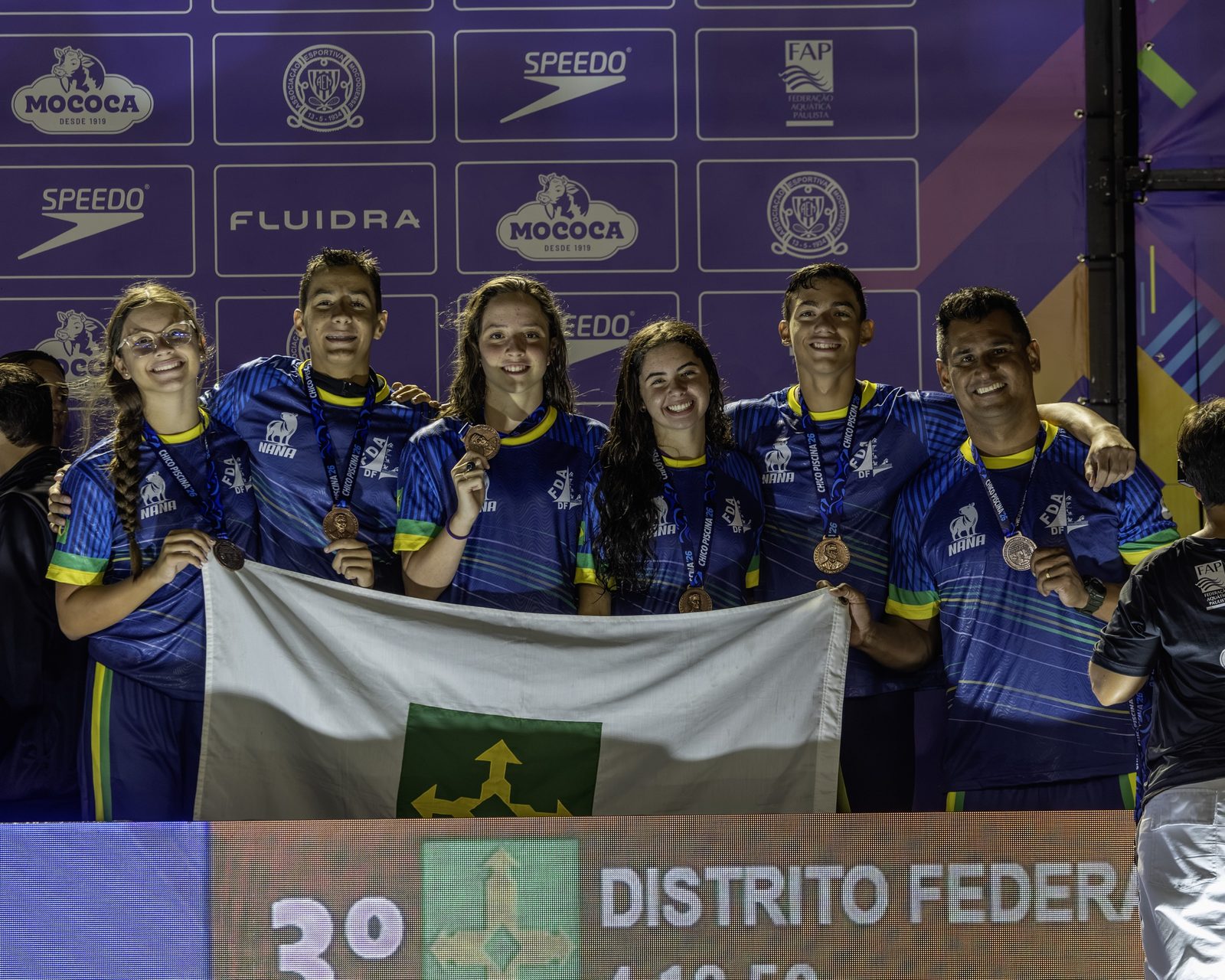 A equipe do DF no pódio do revezamento 4x100 medley misto infantil, com as medalhas de bronze e a bandeira do Distrito Federal