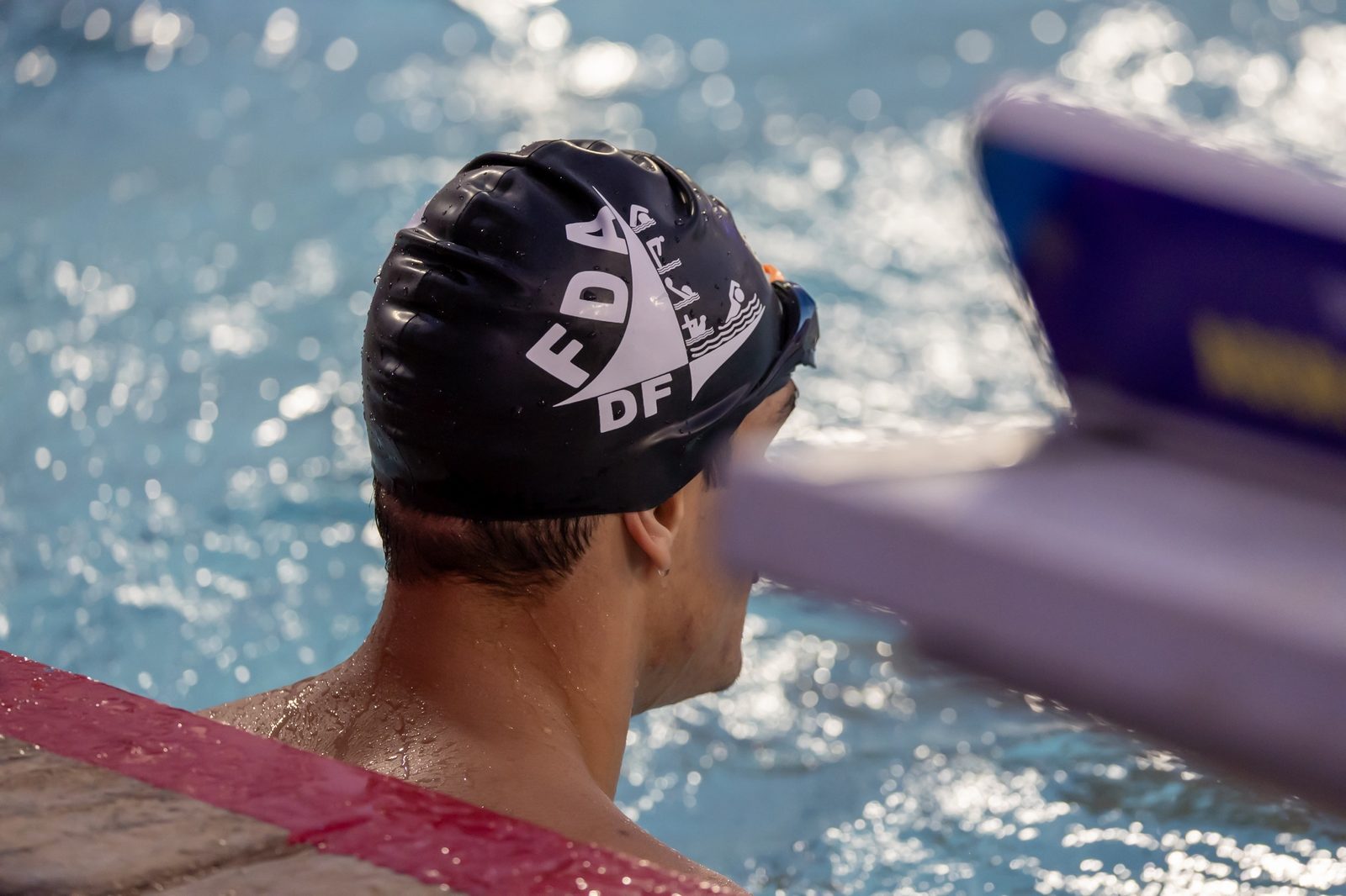 Atleta de touca da FDA-DF, de costas, apoiado na borda da piscina, com a água brilhando ao fundo